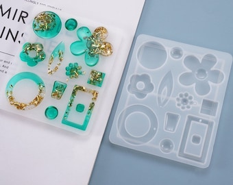 May include: A clear silicone mold with 14 different shapes for making resin jewelry. The shapes include circles, squares, flowers, and leaves. The mold is shown with finished resin pieces in green and gold.