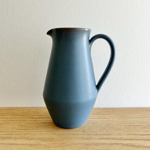 May include: A blue ceramic pitcher with a wide base and a narrow neck. The pitcher has a rounded handle and a smooth, matte finish.