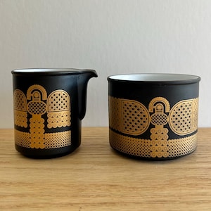 Puede incluir: Un juego de jarra para crema y azucarero negros con un patrón geométrico dorado. La jarra para crema tiene un pico y un asa, mientras que el azucarero es cilíndrico. Ambas piezas tienen un interior blanco y están sobre una superficie de madera.