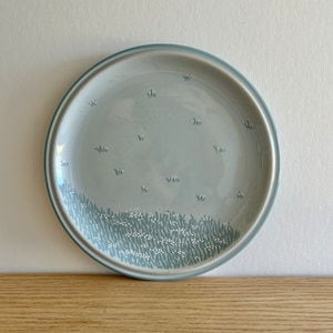 Könnte beinhalten: Ein hellblauer Keramikteller mit einem weißen Blumendesign. Der Teller hat einen weißen Rand und eine hellblaue Mitte mit einem Muster aus weißen Blumen und grünen Blättern.