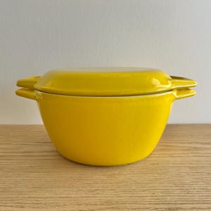 Puede incluir: Una fuente para horno de color amarillo brillante con tapa y asas laterales. La fuente, probablemente de cerámica o hierro fundido esmaltado, está sobre una superficie de madera. La tapa encaja bien y la fuente tiene un acabado brillante.