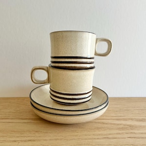 May include: Two beige ceramic mugs with dark brown horizontal stripes are stacked on top of each other. The mugs have rounded handles and sit on a matching saucer. The set is on a wooden surface.