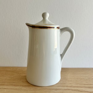 May include: White ceramic creamer with a gold and brown trim around the top. The creamer has a lid and a handle.