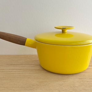 Pode incluir: Uma panela de ferro fundido esmaltado amarelo brilhante com tampa e cabo de madeira. A alça é fixada na lateral da panela. A tampa tem uma pequena alça redonda. A panela está em uma superfície de madeira.