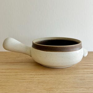 May include: A speckled white ceramic soup bowl with a brown rim and handle. The bowl has a small handle on the side opposite the larger handle. The interior of the bowl is dark brown.