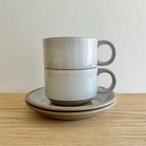 May include: Two stacked, speckled, light gray ceramic mugs with rounded handles, sitting atop two matching saucers. The mugs and saucers have a minimalist design and a neutral color palette, perfect for a modern kitchen.