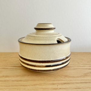 May include: A cream-coloured ceramic sugar bowl with a lid, adorned with dark brown horizontal stripes. The lid has a knob and a small spoon opening. The bowl sits on a wooden surface.
