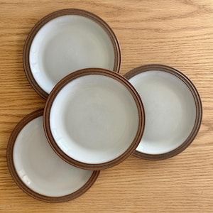 Puede incluir: Cuatro platos de cerámica blanca con borde marrón y una banda marrón alrededor del borde. Los platos están dispuestos sobre una superficie de madera clara. Los platos son redondos y tienen un diseño sencillo y clásico.