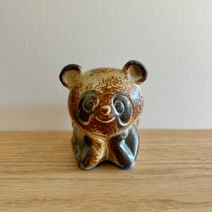 Puede incluir: Una figurita de panda de cerámica marrón y blanca con ojos negros y nariz marrón. El panda está sentado con las patas cruzadas delante de él.