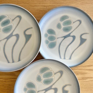 Op de afbeelding: Een set van drie keramische borden met een lichtblauw bloemmotief. De borden hebben een witte achtergrond en een lichtblauwe rand. Het bloemmotief is in blauw- en beigetinten.