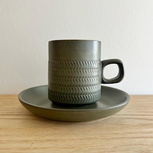 May include: A ceramic mug and saucer set in a muted green hue. The mug features a textured, repeating pattern around its midsection and a rounded handle. The saucer is a simple, shallow dish. The set is displayed on a wooden surface.