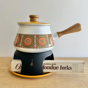 Può includere: Un set per fonduta vintage con una pentola smaltata bianca con un motivo, manico e coperchio in legno. La pentola poggia su un supporto nero con base gialla, e una scatola di forchette per fonduta in acciaio inossidabile è di fronte.