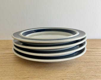 May include: A stack of five white ceramic plates with a dark blue border. The plates are stacked on top of each other, with the top plate showing a full view of the design. The plates are on a light brown wooden surface.