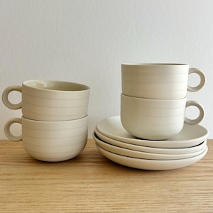 May include: A set of beige ceramic cups and saucers. The cups have a rounded shape with a handle and horizontal lines. The saucers are round and stacked. The items are arranged on a wooden surface against a white background.