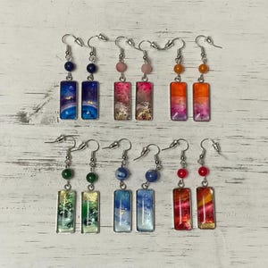 May include: A collection of handmade dangle earrings. Each pair features a rectangular resin pendant with colorful swirls and metallic accents, paired with a beaded accent and silver-tone ear wires. The earrings showcase a variety of colors, including blue, pink, orange, green, and red.