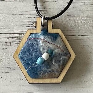 May include: A handcrafted necklace with a hexagonal wooden frame, showcasing a blue and white fibre art design. The pendant is embellished with blue and white beads, and red and white thread details. The necklace is suspended from a black cord.