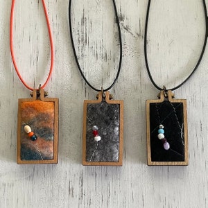May include: Three necklaces with wooden frames and felt inserts. The necklaces are on a white wooden background. The felt inserts are in different colours and patterns. One is orange and brown with white and black beads. One is grey with white and red beads. One is black with white and blue beads.