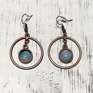 Può includere: Un paio di orecchini a cerchio color rame. Ogni orecchino presenta un grande cerchio circolare con un pendente più piccolo, rotondo, blu e dorato sospeso al centro. Gli orecchini hanno una semplice chiusura a gancio.