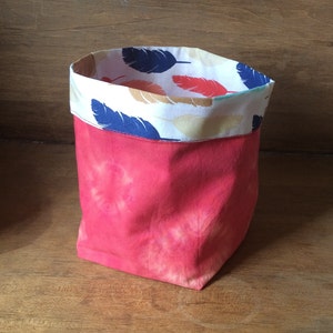 Peut inclure: Un panier de rangement en tissu rouge avec une doublure à motif blanc et bleu représentant des plumes. Le panier est fait d'un tissu doux et texturé et a un effet tie-dye.