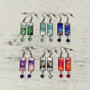 May include: A collection of handmade earrings with rectangular, resin-filled pendants in various colors. Each earring features a unique design with silver-tone hooks and a dangling bead. The color palette includes green, blue, purple, and red.