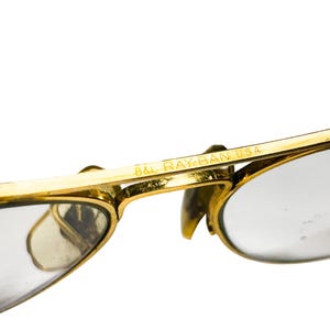 Puede incluir: Primer plano de gafas de sol Ray-Ban doradas vintage. Las monturas son de color dorado brillante, con lentes transparentes. El texto "B&L RAY-BAN USA" es visible en la montura. Dise&ntilde;o cl&aacute;sico y atemporal.