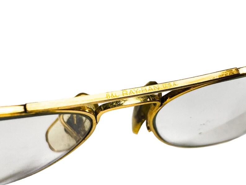 Puede incluir: Primer plano de gafas de sol Ray-Ban doradas vintage. Las monturas son de color dorado brillante, con lentes transparentes. El texto "B&L RAY-BAN USA" es visible en la montura. Dise&ntilde;o cl&aacute;sico y atemporal.