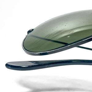 May include: Close-up of a pair of aviator sunglasses with a dark green tint. The lenses are slightly scratched. The frames and arms are a dark, glossy color. The sunglasses are set against a white background.