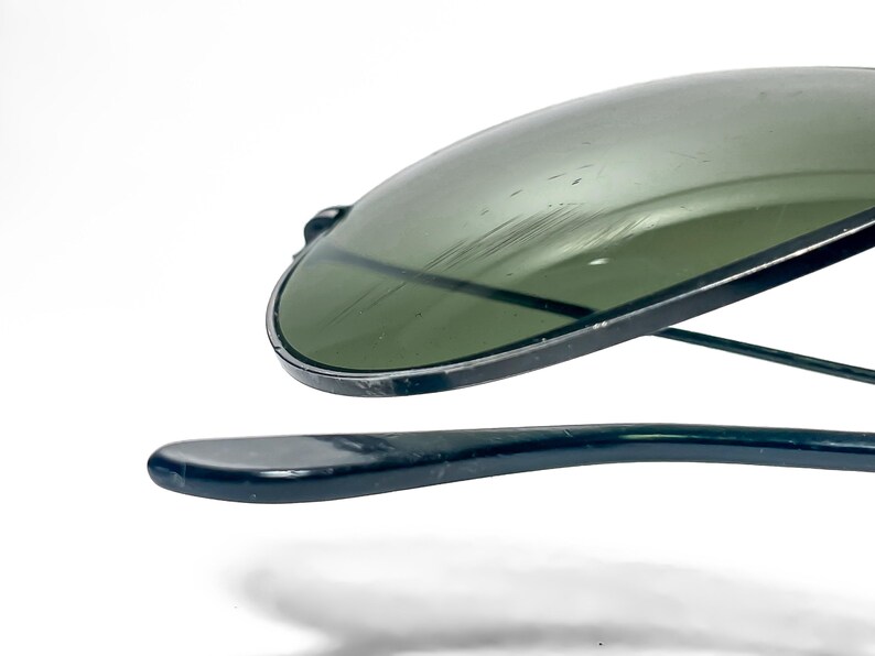 May include: Close-up of a pair of aviator sunglasses with a dark green tint. The lenses are slightly scratched. The frames and arms are a dark, glossy color. The sunglasses are set against a white background.