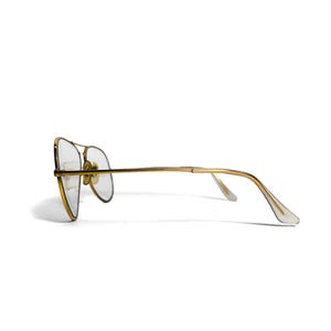 Puede incluir: Gafas de aviador con montura dorada y lentes transparentes. Las gafas tienen un doble puente y un dise&ntilde;o cl&aacute;sico. Las patillas son finas y se extienden hasta los terminales transparentes. Las gafas est&aacute;n sobre fondo blanco.