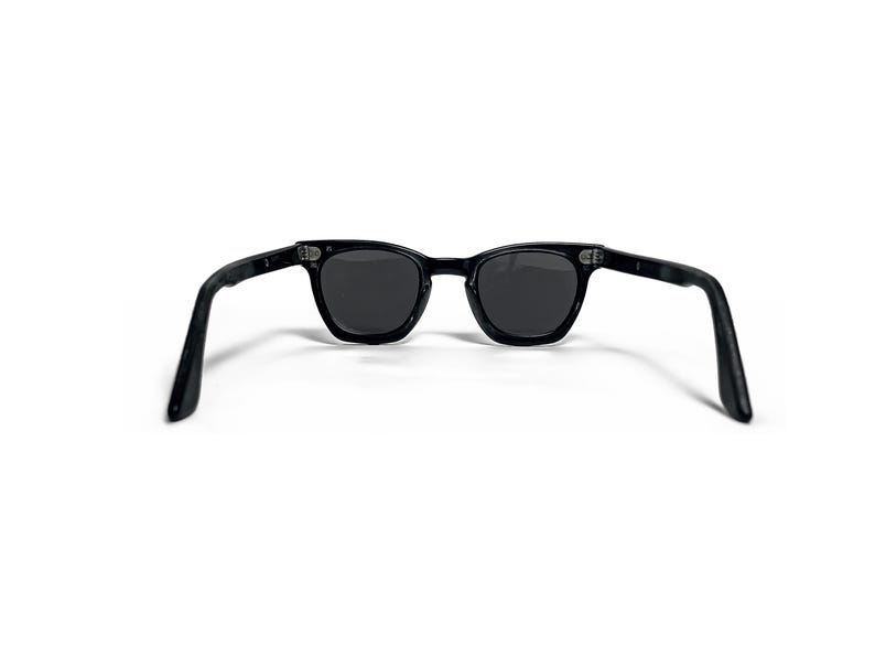 May include: Black sunglasses with dark lenses. The sunglasses have a classic, rectangular frame and are set against a white background. The arms of the sunglasses are extended, showing the full shape.