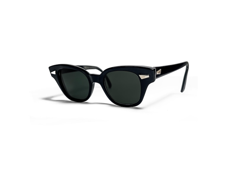 May include: Black sunglasses with a cat-eye shape and dark lenses. The frames are a glossy black with silver-colored accents on the temples. The sunglasses are set against a white background.