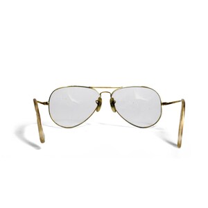 Puede incluir: Gafas de sol estilo aviador con montura de metal dorado y lentes transparentes. Las lentes tienen forma de l&aacute;grima. Las patillas son de color beige claro. Las gafas de sol est&aacute;n sobre un fondo blanco.