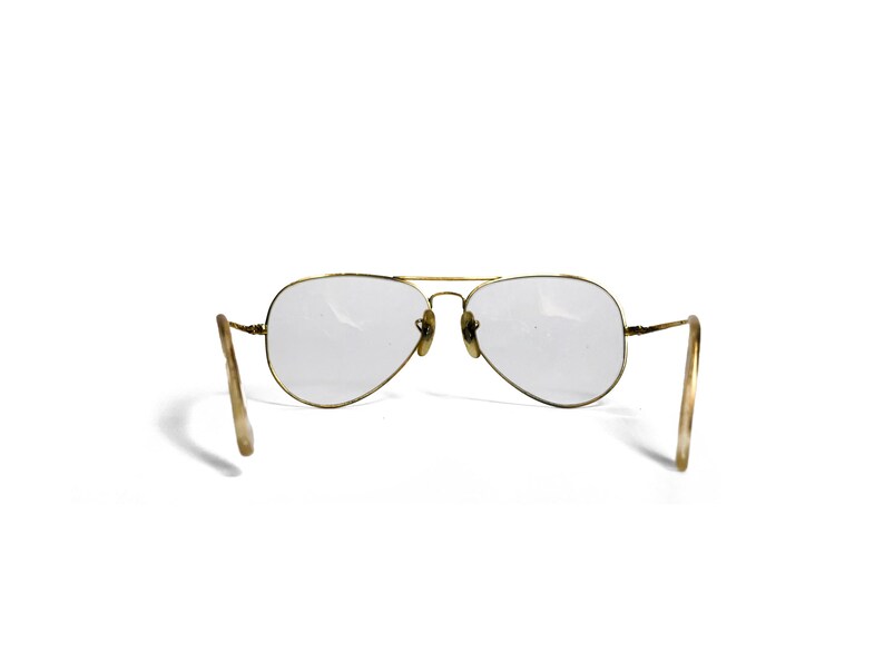 Puede incluir: Gafas de sol estilo aviador con montura de metal dorado y lentes transparentes. Las lentes tienen forma de l&aacute;grima. Las patillas son de color beige claro. Las gafas de sol est&aacute;n sobre un fondo blanco.