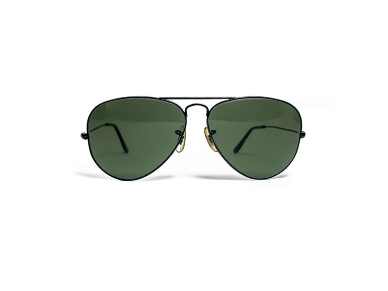 May include: Aviator-style sunglasses with a black metal frame and green lenses. The sunglasses have a double bridge and gold-colored accents on the hinges. The lenses are a teardrop shape, and the arms are thin and black.