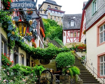 May include: Picturesque European village scene with stone buildings, stairs, and lush greenery. A sign reading "Winzerstube" hangs above a lantern. Flowers and vines adorn the buildings, creating a charming, inviting atmosphere. Outdoor seating is visible.