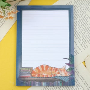 May include: A blue lined notepad with a cat sleeping on a bookshelf with books. The cat is orange and white with a black nose. The notepad has a blue border.