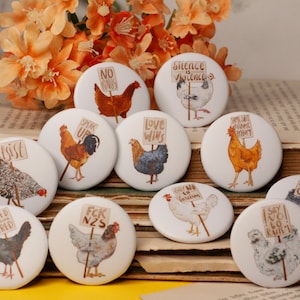 May include: A selection of white circular badges with chicken illustrations, each holding a sign with slogans such as "No Kings" and "Love Wins." The badges are displayed on a pile of vintage books, with orange flowers in the background.