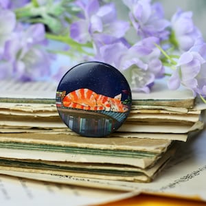 May include: A round button with a dark blue background featuring a sleeping orange tabby cat on a rooftop with books. The button is set on a stack of old book pages, with purple flowers in the background.