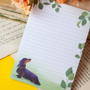 Peut inclure: Bloc-notes ligné avec une illustration de teckel. Le bloc-notes a une bordure verte avec des accents de feuilles. Le teckel est noir et marron avec un collier rouge. Le fond comprend de vieux livres et des fleurs.
