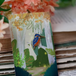 Op de afbeelding: Kleurrijke emaille pin met een blauwe, oranje en witte vogel op een tak. De pin staat tegen een boslandschap met de tekst "Tiny Little Forest". Decoratief accessoire.