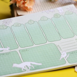 May include: A green weekly planner with a grid layout and illustrations of animals. The planner has sections for Monday, Tuesday, Wednesday, Thursday, Friday, Saturday, and Sunday, as well as sections for notes, habits, and next week.
