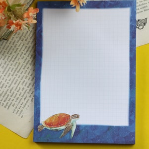 Peut inclure: Un bloc-notes quadrillé bleu et blanc avec une illustration de tortue de mer en bas. Le bloc-notes a une bordure bleue avec un motif de nuit étoilée.
