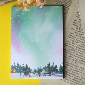 May include: A snowy winter scene with a small red cabin and two reindeer in the foreground. The sky is a vibrant green and purple with a hint of pink, suggesting the aurora borealis. The trees are silhouetted against the colourful sky.