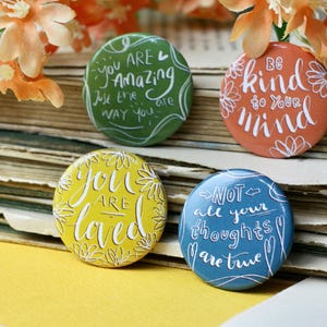 Könnte beinhalten: Vier bunte runde Buttons mit inspirierenden Zitaten. Die Buttons sind grün, orange, gelb und blau. Der grüne Button sagt: "You are Amazing, Just the Way You Are". Der orangefarbene Button sagt: "Be Kind to Your Mind."
