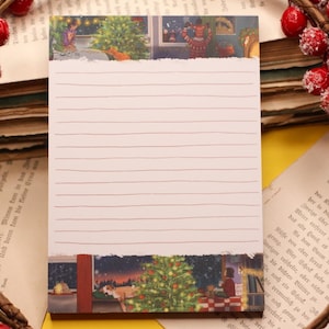 Könnte beinhalten: Ein weißer Notizblock mit liniertem Papier und einer Weihnachtsszene oben und unten. Die obere Szene zeigt eine Familie, die um einen Weihnachtsbaum versammelt ist, während die untere Szene eine verschneite Winterlandschaft mit einem Haus und einem Weihnachtsbaum zeigt.