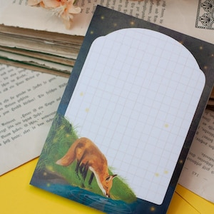 May include: A decorative notepad featuring a fox drinking from a stream. The notepad has a white grid pattern for writing, with a dark blue border and a starry night sky design. The notepad is placed on a yellow surface with old books.