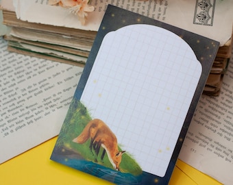 May include: A decorative notepad featuring a fox drinking from a stream. The notepad has a white grid pattern for writing, with a dark blue border and a starry night sky design. The notepad is placed on a yellow surface with old books.