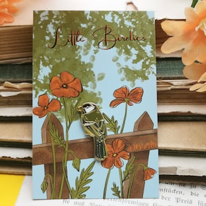 May include: Gold enamel pin featuring a small bird perched on a fence with a blue sky and orange flowers in the background. The text "Little Birdies" is at the top of the card.