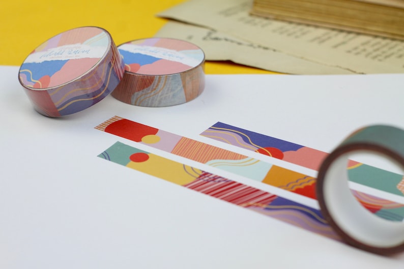 May include: Three rolls of colorful patterned washi tape with abstract designs. The tape is unrolled on a white surface, showing the different patterns. The tape is labeled "Gold Star".