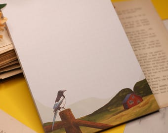 Puede incluir: Bloc de notas con una ilustración en acuarela de una urraca posada en un poste de madera, con una casa roja en una ladera al fondo. El bloc de notas está rodeado de libros antiguos y flores.